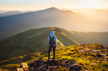 Wall Mural - Girl on mountain peak with green grass looking at beautiful mountain valley in fog at sunset in summer. Landscape with young woman on the trail, foggy hills, forest, sky. Travel and tourism. Hiking	