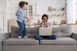 © DimaBerlin - Irritated mother sitting on couch working on laptop at home, distracted from noise and touching her temple, hyperactive little kid jumping on sofa and drawing attention. Tired mom can't concentrate.