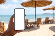 Mockup image of a man holding mobile phone with blank desktop screen while sitting on the beach