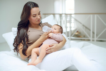 Smiling mother feeding baby boy with fresh milk in plastic bottle in bed closeup. Looking at camera. Motherhood. Childhood. High quality photo.