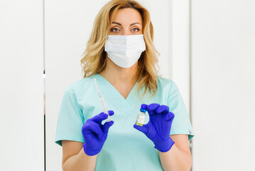 Young attractive blondy doctor in blue medical coat, mask and blue gloves posing with syringe and  vaccine against white background.