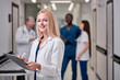 © Roman - Pretty Female Doctor In White medical Unifrom with clipboard in hospital, standing in aisle, taking notes, checking information about patients, smile at camera