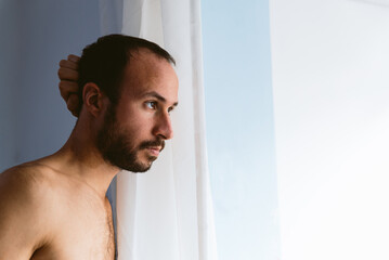 young adult pensive man in his 30s looking out of window with serious and confident expression. simple natural light. candid moment with copy space