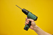 Green Cordless Battery Powered Drill On Yellow Background, Cropped Male Hands Holding Tool For Repair And Building Construction. Copy Space For Advertisement