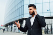 © Тарас Нагирняк - A portrait of an Indian man, an office worker, a bank employee talking on wireless headphones and gesturing, behind him a large modern building