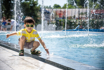 Wall Mural - Little boy plays in the square near pool with water jets in the fountain at sunny summer day. Active summer leisure for kids in the city