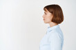 © Cookie Studio - Side portrait of young female entrepreneur with short hair, wearing office blouse, looking left at copy space with serious face, standing on white background
