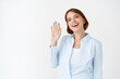 © Cookie Studio - Portrait of happy friendly woman saying hello, waving hand hi greeting gesture, smiling at camera, standing in office blouse on white background