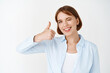 © Cookie Studio - Well done. Smiling successful businesswoman winking, say yes and showing thumb up in approval, like something good, recommending product, standing on white background