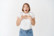 © Cookie Studio - Shocked young woman with short hair, gasping wondered, open mouth and holding hands pressed to chest, standing on white background