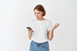 © Cookie Studio - Confused young woman looking at mobile phone with questioned expression, shrugging troubled, standing against white background