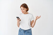 © Cookie Studio - Annoyed girl look at smartphone screen and complaining, staring angry at display with raised hand, standing against white background