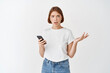 © Cookie Studio - Confused girl dont understand message, look puzzled and shrugging after reading smartphone screen, standing against white background