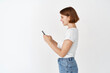 © Cookie Studio - Profile of young woman chatting on mobile phone. Happy girl with short hair texting message with smartphone, standing against white background