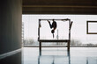 © BASILICOSTUDIO STOCK - Long shot of a big Pilates Studio with wide windows and outside views, with a fit woman hanging upside down from a Cadillac Machine, Trapeze Table. Pilates instructor. Back lit, shadows.