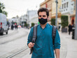 © David Martínez - Hombre joven con mochila caminando por la calle vistiendo una mascarilla negra cumpliendo las medidas de seguridad y distanciamiento social de la pandemia del coronavirus