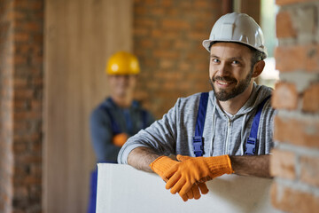 portrait of positive, handsome young male builder in hard hat smiling at camera, holding drywall whi