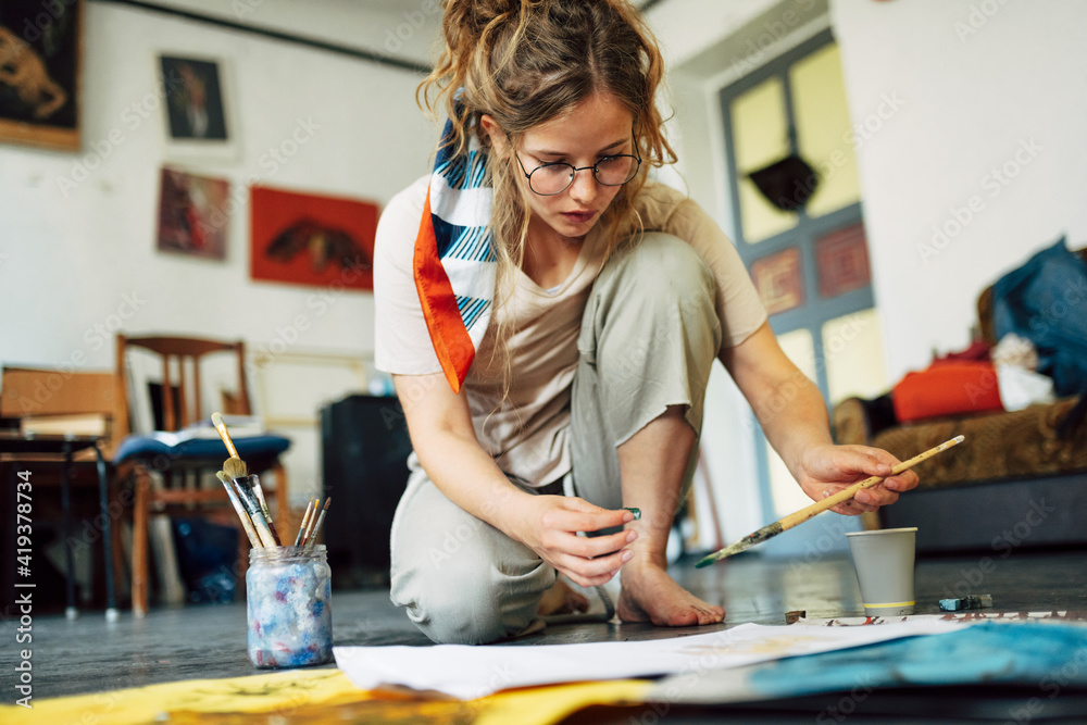 Horizontal image of a pretty female artist sitting on the floor in the art studio and painting on paper with a brush. A woman painter with glasses painting with watercolors in the workshop. - obrazy, fototapety, plakaty 