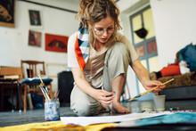Horizontal Image Of A Pretty Female Artist Sitting On The Floor In The Art Studio And Painting On Paper With A Brush. A Woman Painter With Glasses Painting With Watercolors In The Workshop.