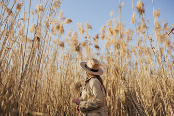 A woman in the beige trench coat and hat posing in the pampas grass with blue sky on background.