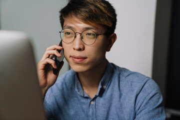 Canvas Print - Serious asian guy talking on cellphone while working with laptop