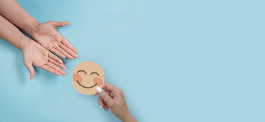 Wall Mural - Hand holding nude paper cut happy smile face give it to another person on blue background, positive thinking, mental health assessment , world mental health day concept