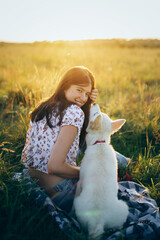 Wall Mural - Happy woman cuddling with cute white puppy in summer meadow in  sunset. Happiness. Summer vacation