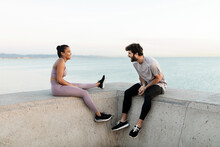 Cheerful Young Bearded Sportsman Speaking With Content Ethnic Female Athlete While Resting Against Endless Sea And Looking At Each Other
