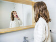 © Albert Morcillo/ADDICTIVE STOCK - Side view happy little girl wearing cozy white sweater looking in mirror with toothy smile while standing with toothbrush in modern bathroom