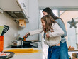© Albert Morcillo/ADDICTIVE STOCK - Cheerful young mother lifting cute smiling daughter stirring food on frying pan while cooking together in contemporary kitchen
