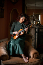 Female Musician Holding Violin While Sitting On Armchair In Vintage Styled Room During Rehearsal