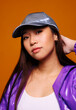 © Victor Torres/ADDICTIVE STOCK - Portrait of Asian young woman with serious expression. She wears a purple jacket and a gray cap and is looking at the camera against a yellow background while touching her head.