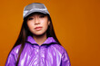 © Victor Torres/ADDICTIVE STOCK - Portrait of Asian young woman with serious expression. She wears a purple jacket and a gray cap and is looking at the camera against a yellow background