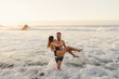 © Carlos Pintau/ADDICTIVE STOCK - Full length of cheerful young man carrying girlfriend and walking out of waving sea water while spending sunset time together on beach of Tenerife island