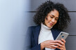 © David Munoz/ADDICTIVE STOCK - Positive young curly haired ethnic businesswoman reading information on mobile phone while standing near gray wall