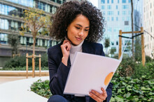 Content Young Ethnic Female Entrepreneur In Stylish Formal Suit Studying Documents In Folder While Sitting On City Street
