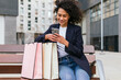 © David Munoz/ADDICTIVE STOCK - Positive black female sitting on bench with paper bags after shopping and browsing mobile phone in city