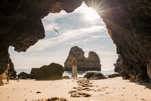 Back View Of Unrecognizable Female In Casual Clothes And Hat Standing On Sand In Entrance Of Cave Near Sea Looking At A Bird In Algarve, Portugal