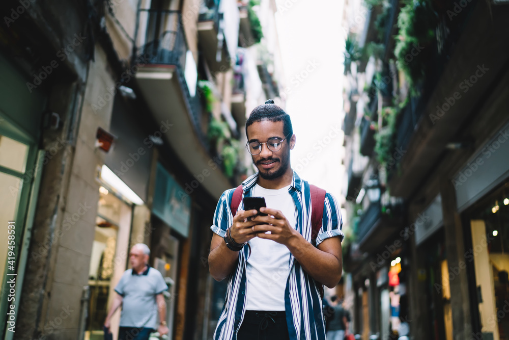 Positive young ethnic man browsing cellphone while walking on street - obrazy, fototapety, plakaty 