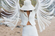 © Erika Anes/ADDICTIVE STOCK - Unrecognizable slim female model in elegant white hat and suit with straps looking like wings raising arms while standing on sandy beach