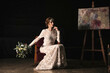 © German Linares/ADDICTIVE STOCK - Full body of beautiful young woman in bohemian style bridal gown with bouquet of flowers sitting on chair near easels with pictures in dark studio