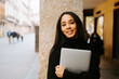 © Jake Jakab/ADDICTIVE STOCK - Charming young ethnic female with netbook smiling at camera while standing at stone column on city street