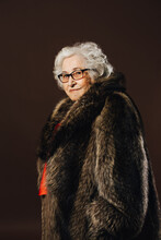 Confident Senior Female In Trendy Fur Coat Standing With Rollator Walker On Brown Background In Studio And Looking At Camera