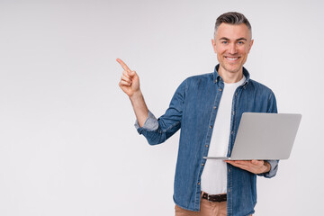 Confident middle-aged man pointing at copy space while holding laptop isolated over white background