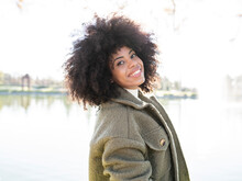 Cheerful Young African American Female With Curly Hair In Stylish Warm Outfit Smiling And Looking At Camera While Resting In Park At Lakeside On Sunny Autumn Day