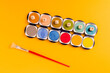 © Jose Lax/ADDICTIVE STOCK - Top view composition with paintbrush placed near colorful palette of watercolor paints with small cubes forming word Colour on yellow background