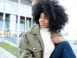 © Jose Carlos Cerdeno/ADDICTIVE STOCK - Trendy young black female millennial with Afro hair in stylish warm clothes resting on street and looking away pensively near modern building with glass walls