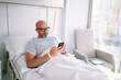 © Jose Luis CARRASCOSA/ADDICTIVE STOCK - Concentrated adult male wearing patient gown and eyeglasses browsing phone on bed in light ward in modern hospital