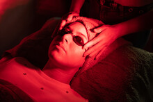 From Above Of Crop Anonymous Female Cosmetologist Adjusting Protective Goggles Of Relaxed Young Woman Lying On Couch During Red LED Light Facial Therapy In Professional Salon
