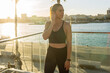 © Marina Gallardo/ADDICTIVE STOCK - Positive fit female in sportswear adjusting wireless earbuds while preparing for outdoor fitness training on city quay at sunset time